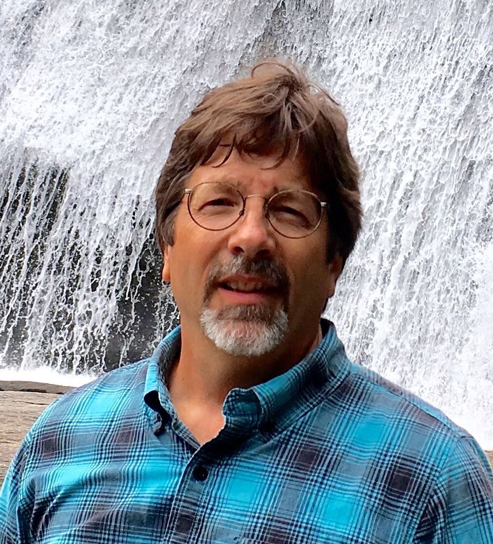 Middle aged man with steel framed classes, brown short hair, and a goatee, standing in front of a waterfall