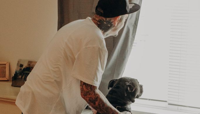 Man with tattoos pets his dog while looking out the window of his CHN apartment