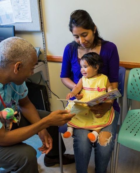 A baby on her mother's lap smiles at a healthare provider while her mother holds a book.