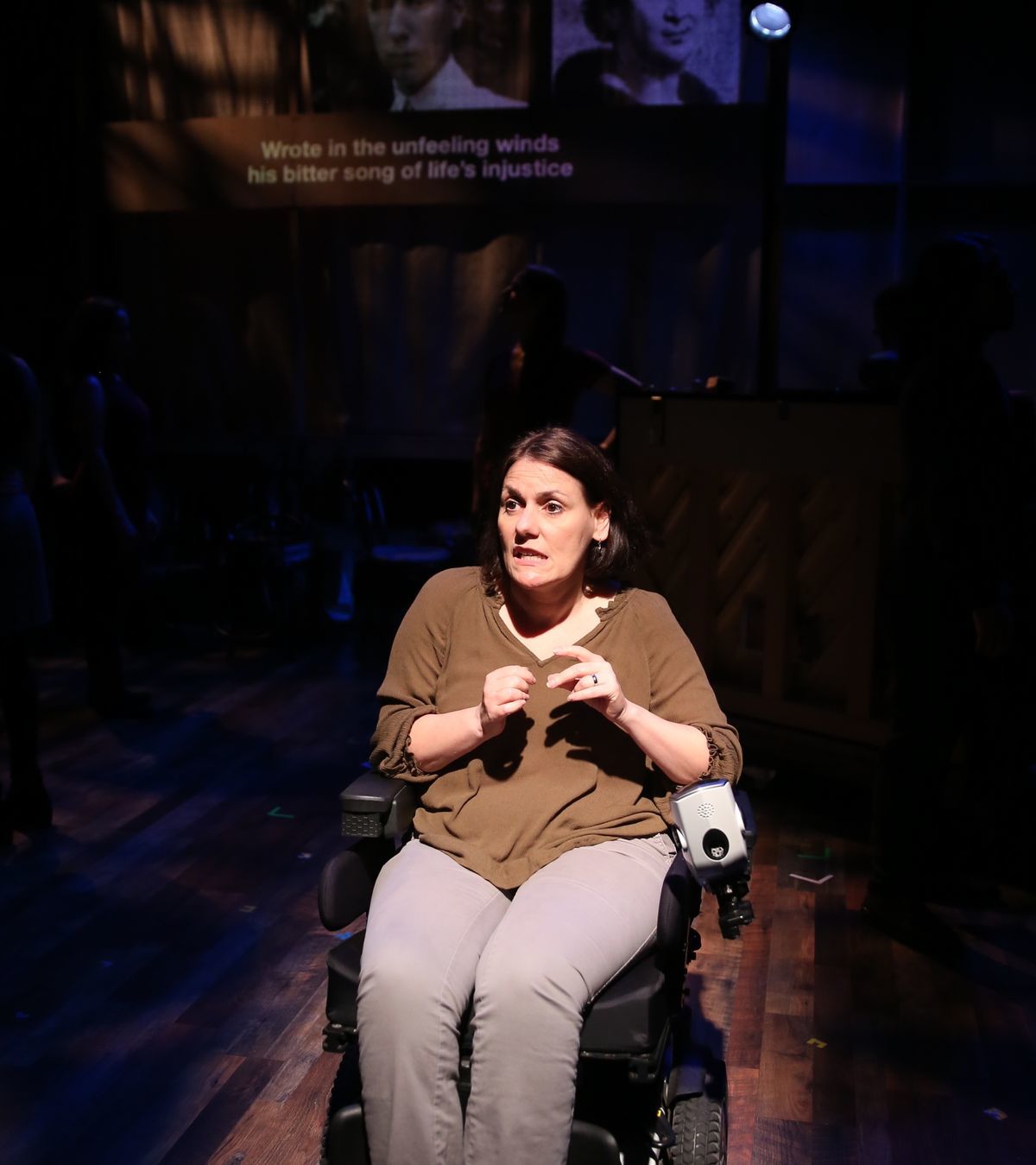 A picture of Ann Marie who’s sitting in her power wheelchair and wearing a green olive shirt and beige pants. She has a spotlight on her and she looks like she is perplexed.