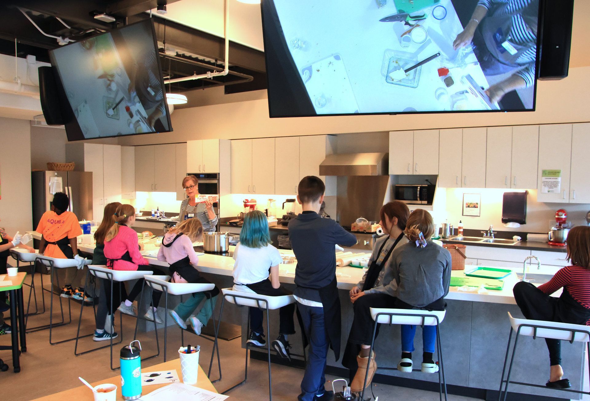 Inside the demonstration kitchen, a teacher leads a class of young students in a cooking lesson.