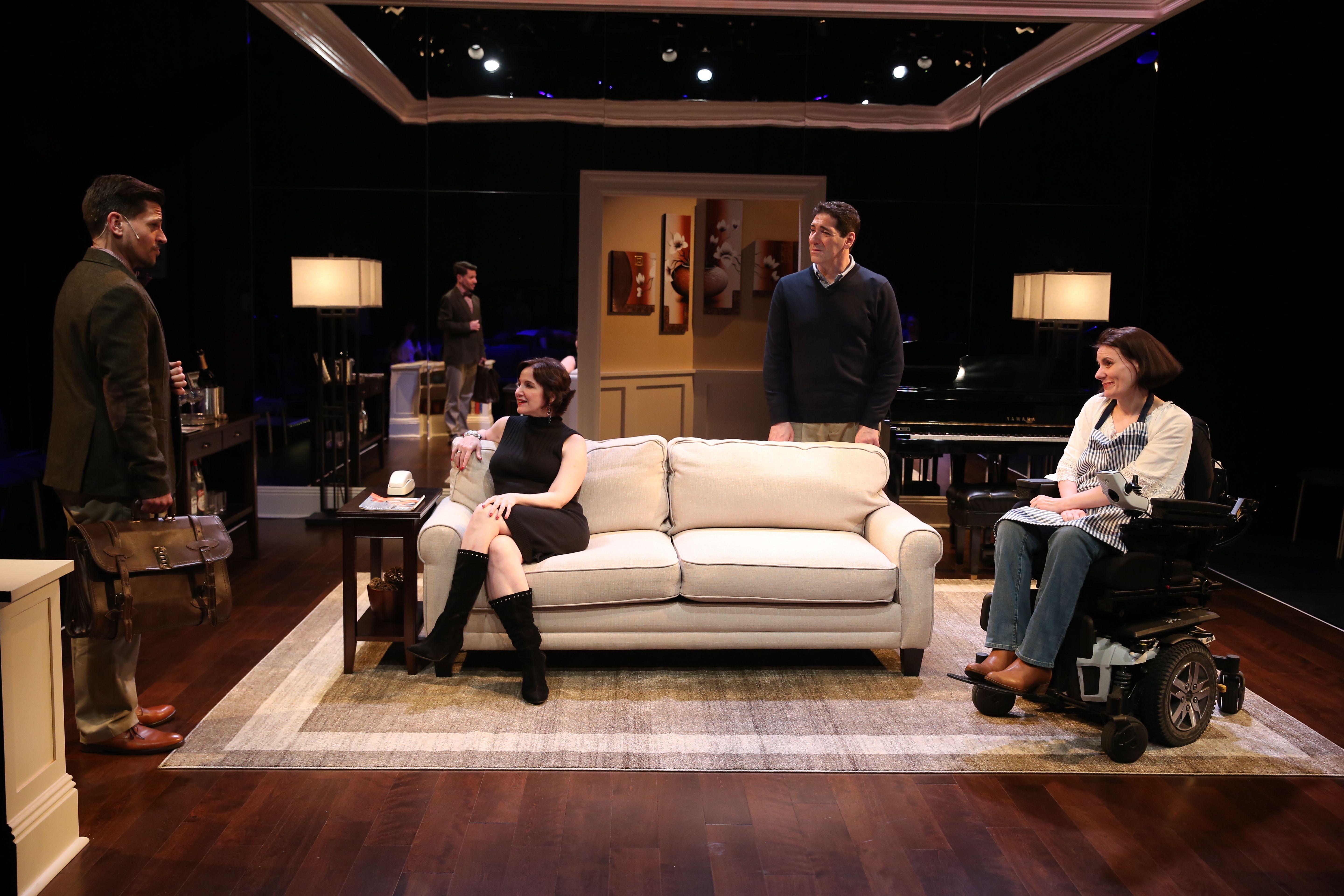 A group shot of actors who are both standing and sitting in a living room. They’re performing a scene from the production of The Fourth Wall. Everyone is looking at an actor who’s standing on the left side of the picture. 