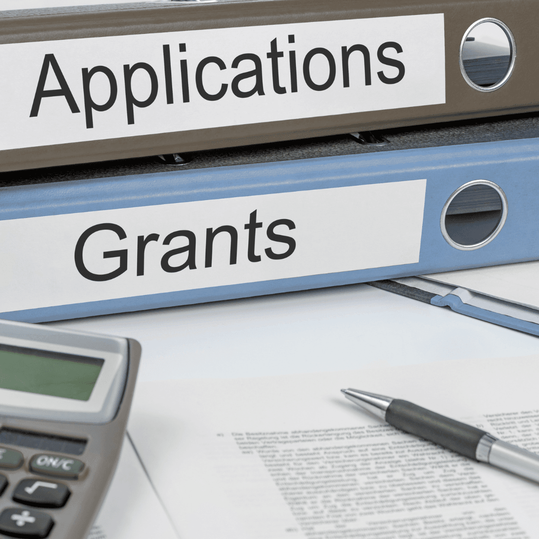 Photo featuring binders labeled with "applications" and grants" with paper, pen, and calculator