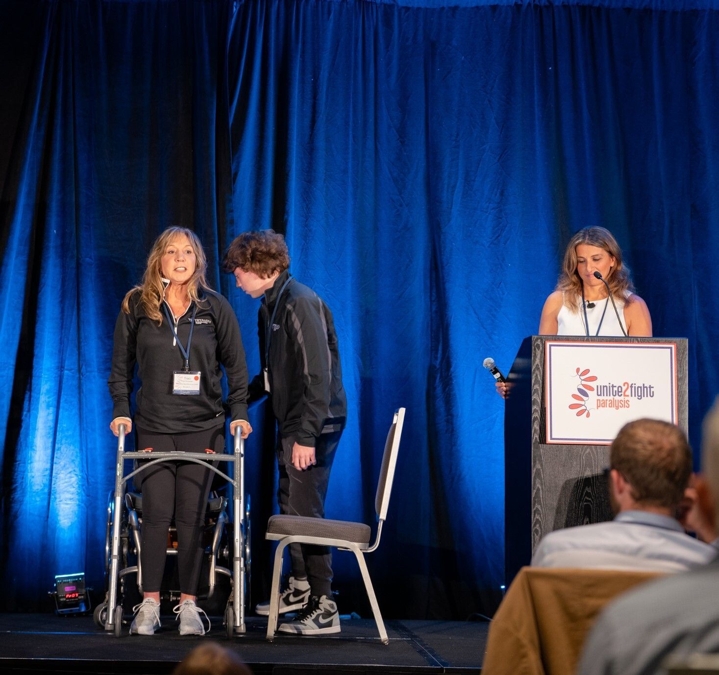 White woman stands with a walker next to a younger white man getting up from a chair while a white woman looks on from behind a lectern