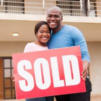 Photo of a young woman and man holding a "Sold" sign in front of their new home. The woman wears a light pink shirt, the man a blue shirt, and the sign has a red background with white letters.