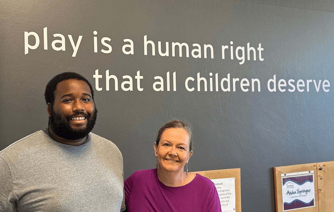 Young man and older woman in purple t-shirt posing for photo in front of quote painted on wall that says "Play is a human right that all children deserve."