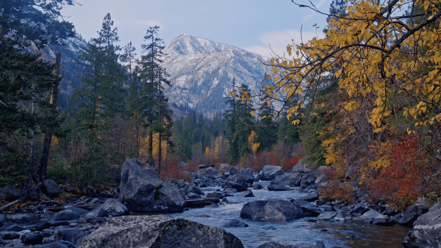 Photo of Icicle Creek running down the middle of a steep valley with a snowy mountain in the background. Surrounding trees are red, green, and yellow and many boulders are scattered in the creek. 
