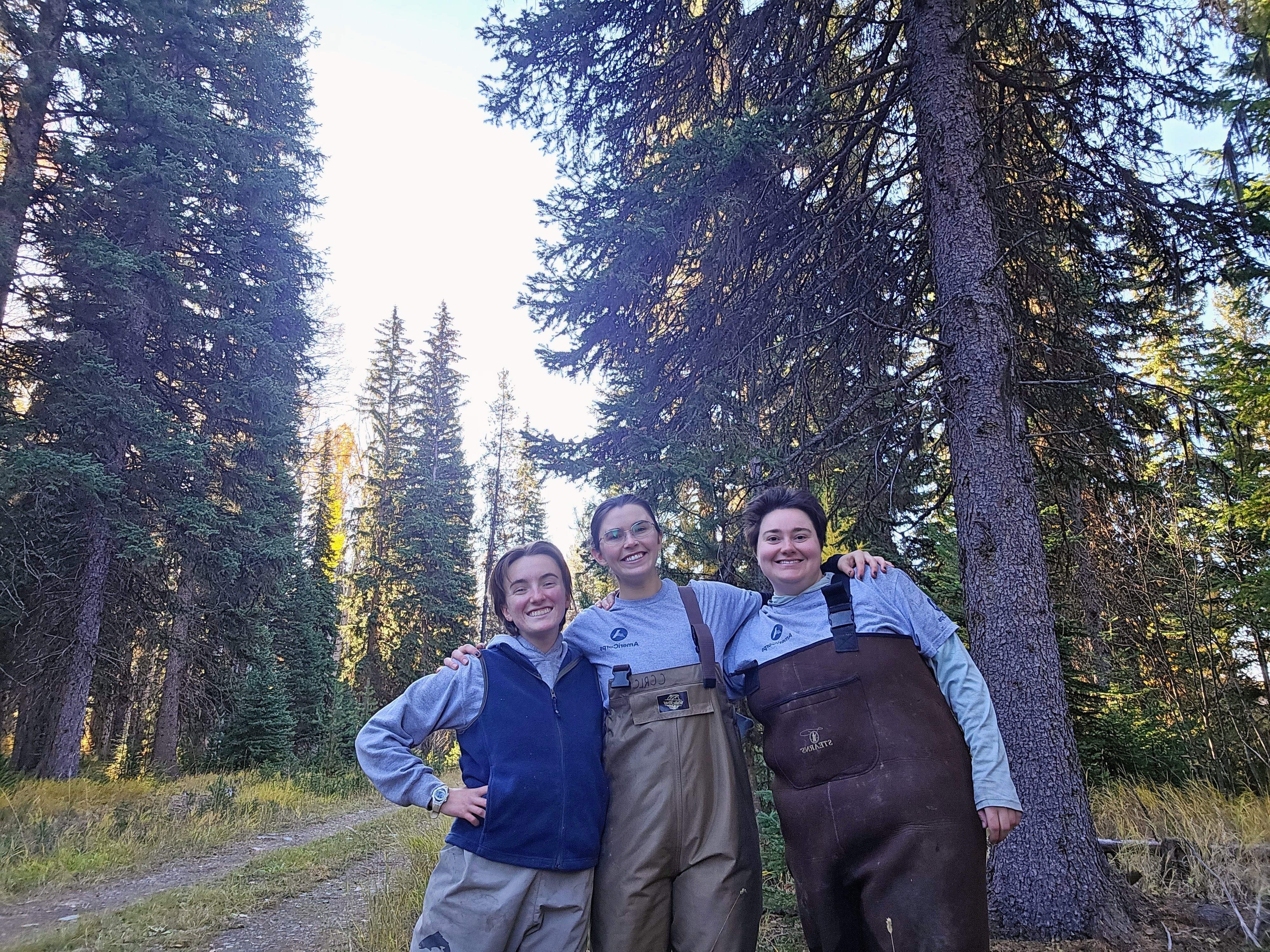 Three MCC members stand wearing gaiters and smiling at the camera during some field work among the woods.