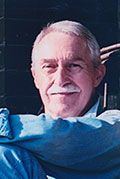 Lawrence Merritt, a man who is in his 60's-70's. He has white hair and a mustache, he is wearing a blue button up shirt, looking towards the camera and crossing his arms, smiling.
