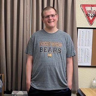 Jason wears a UNC Bears t-shirt and has his hands in his pockets. He stands in his dorm room, smiling.