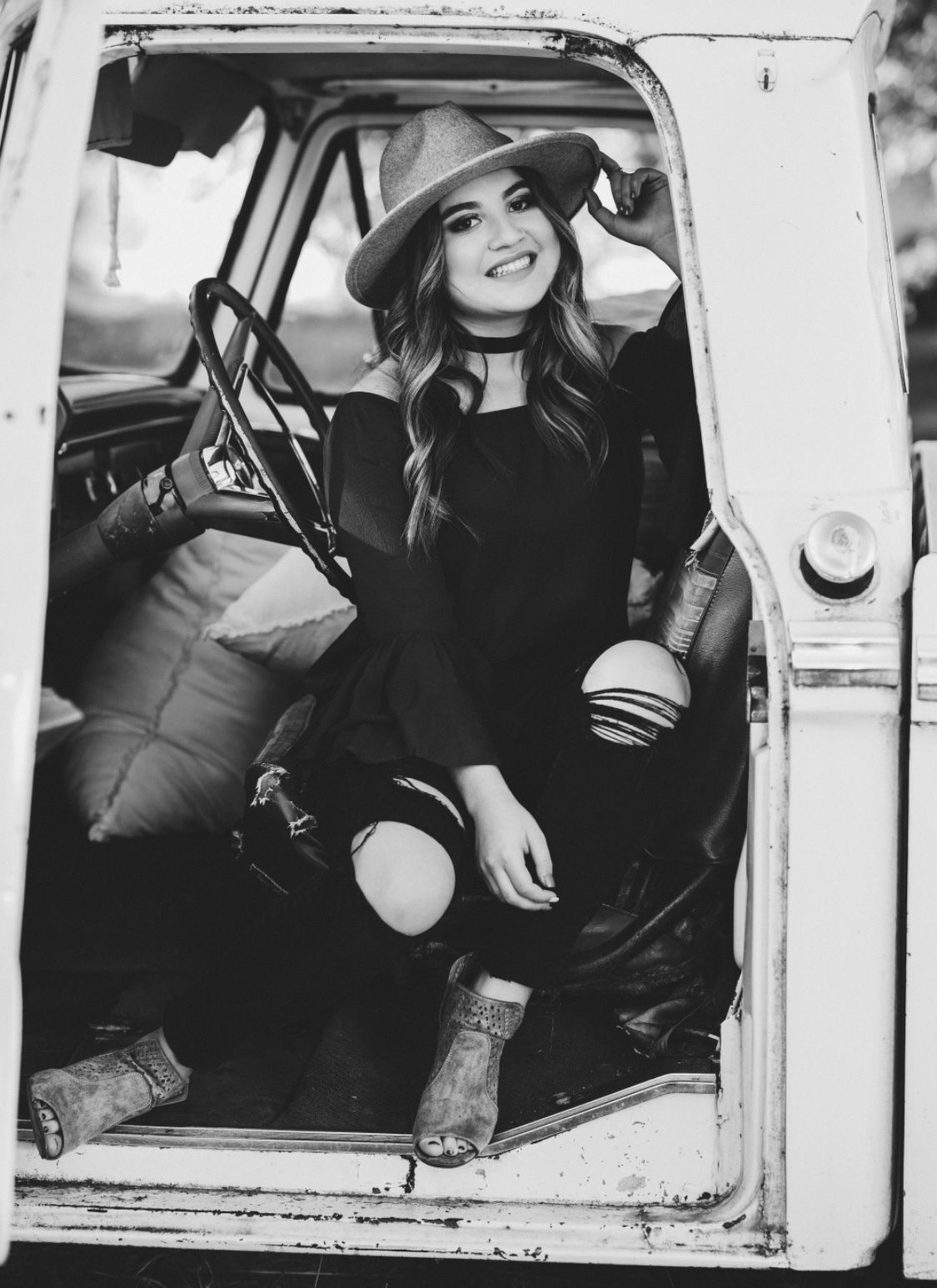 Black and white photo shows a young woman smiling from inside the front seat of what looks like the front of an old truck. She is wearing a wide-brimmed hat, choker, and dark top and pants with open toed boots. 