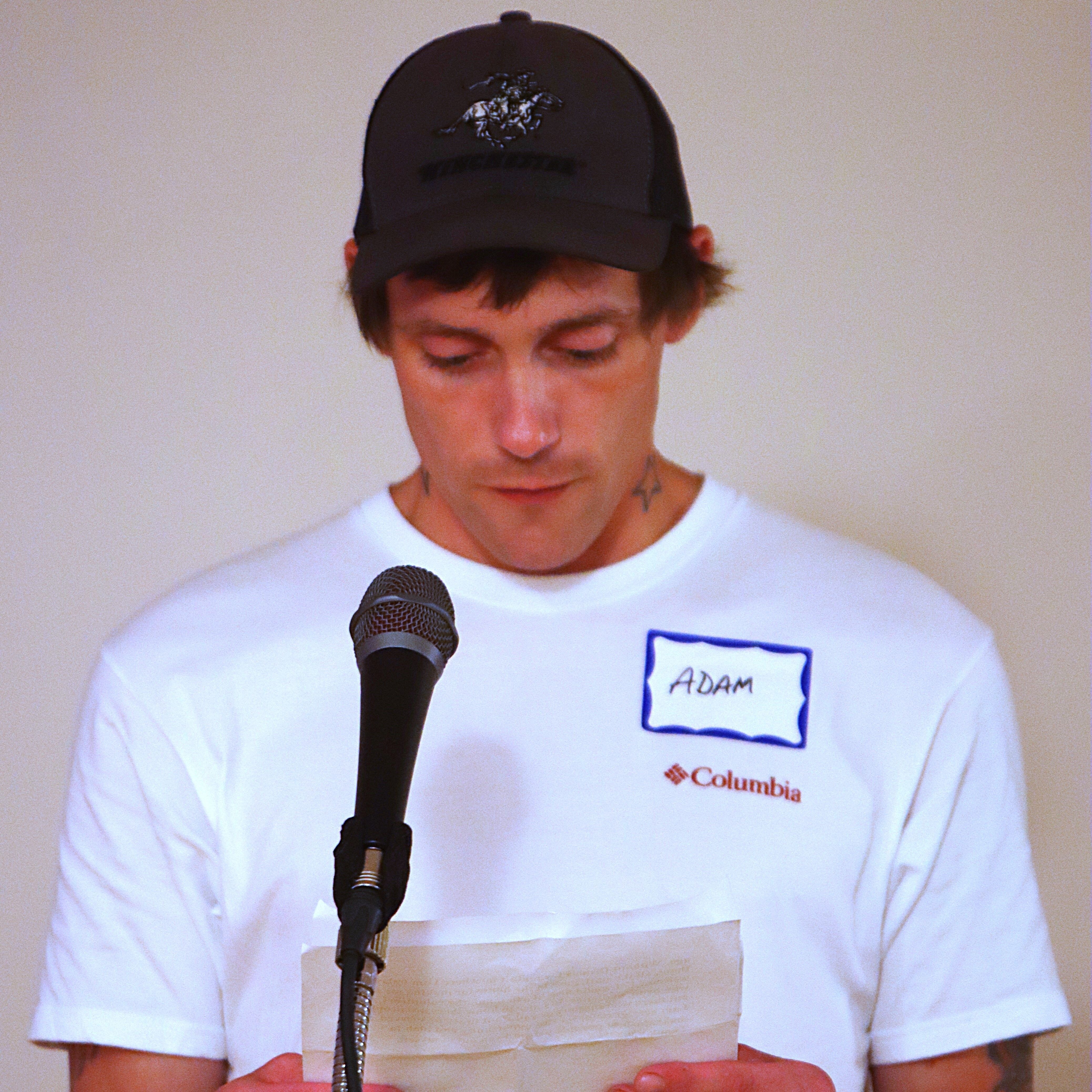 Adam wears a black baseball cap and a white t-shirt with a nametag, he is standing at a microphone and reading from a speech in his hands.