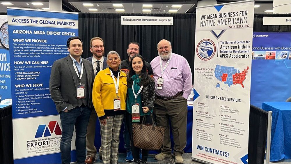 A group of six people at a conference standing between banners for Arizona MBDA Export Center and National Center for American Indian Enterprise Development.