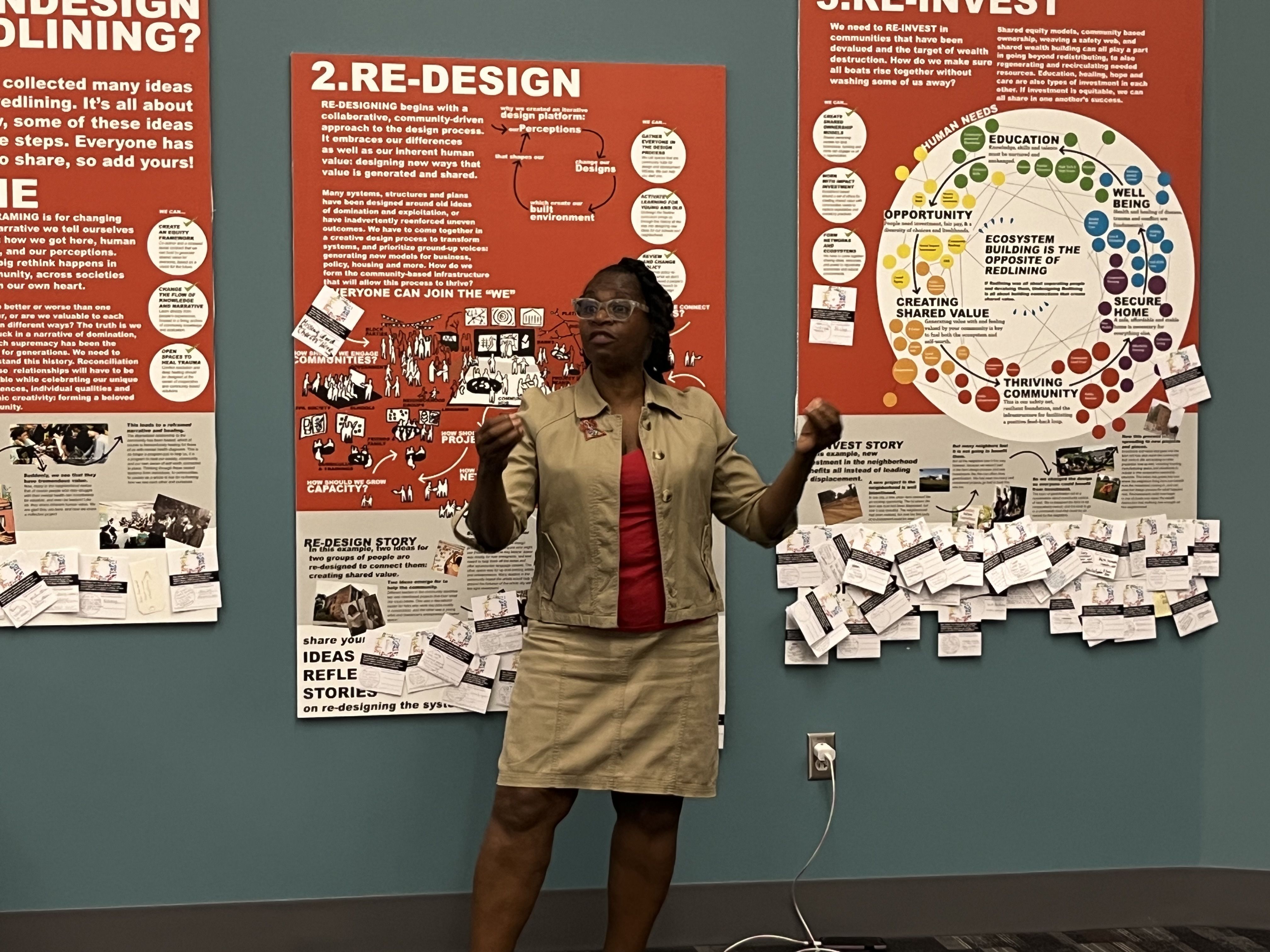 Dr. Terri L. Crawford stands in front of large Undesign the Redline tour photos which are on the wall. She is wearing a beige suit with a red shirt.