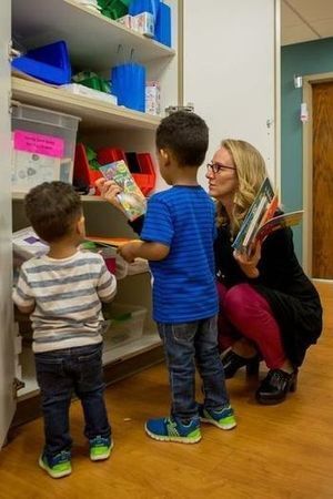 An adult shows two young children books on a bookshelf.