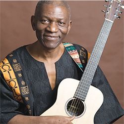 Kenn Wanaku smiling holding his acoustic guitar wearing African patterned sash