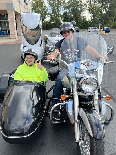 Someone sits in a motorcycle side car facing the camera and giving a "thumbs up" sign while the motorcycle driver faces the camera smiling.