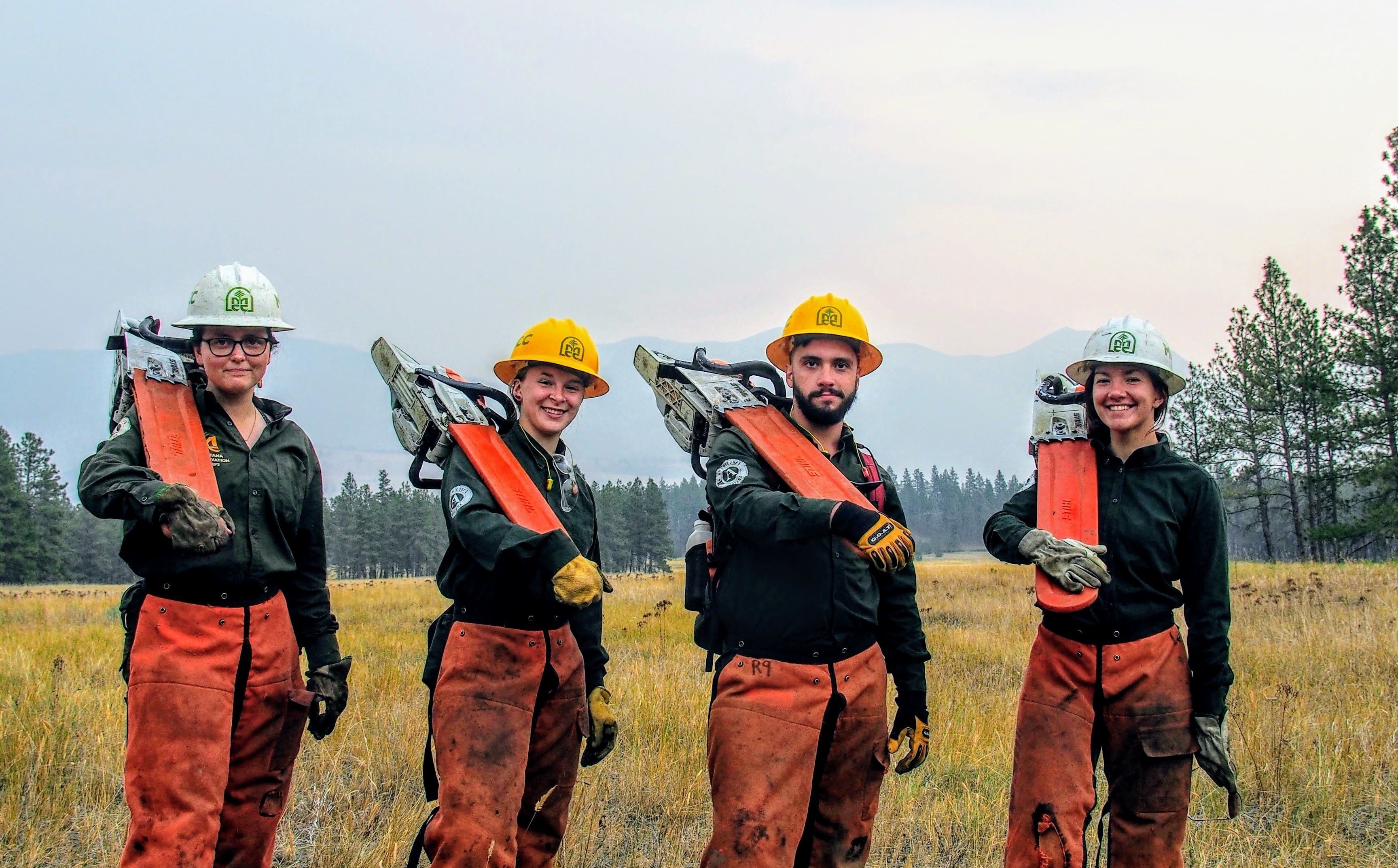 [Image description: Four MCC members holding their chainsaws (with the safety guard on) over their shoulders. In the background, one can see a smoky pink sky.]