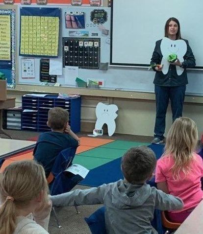 A woman with long hair is holding a large paper cutout of a tooth. She is standing in a classroom and there are young children are sitting on the floor and looking at the woman. 