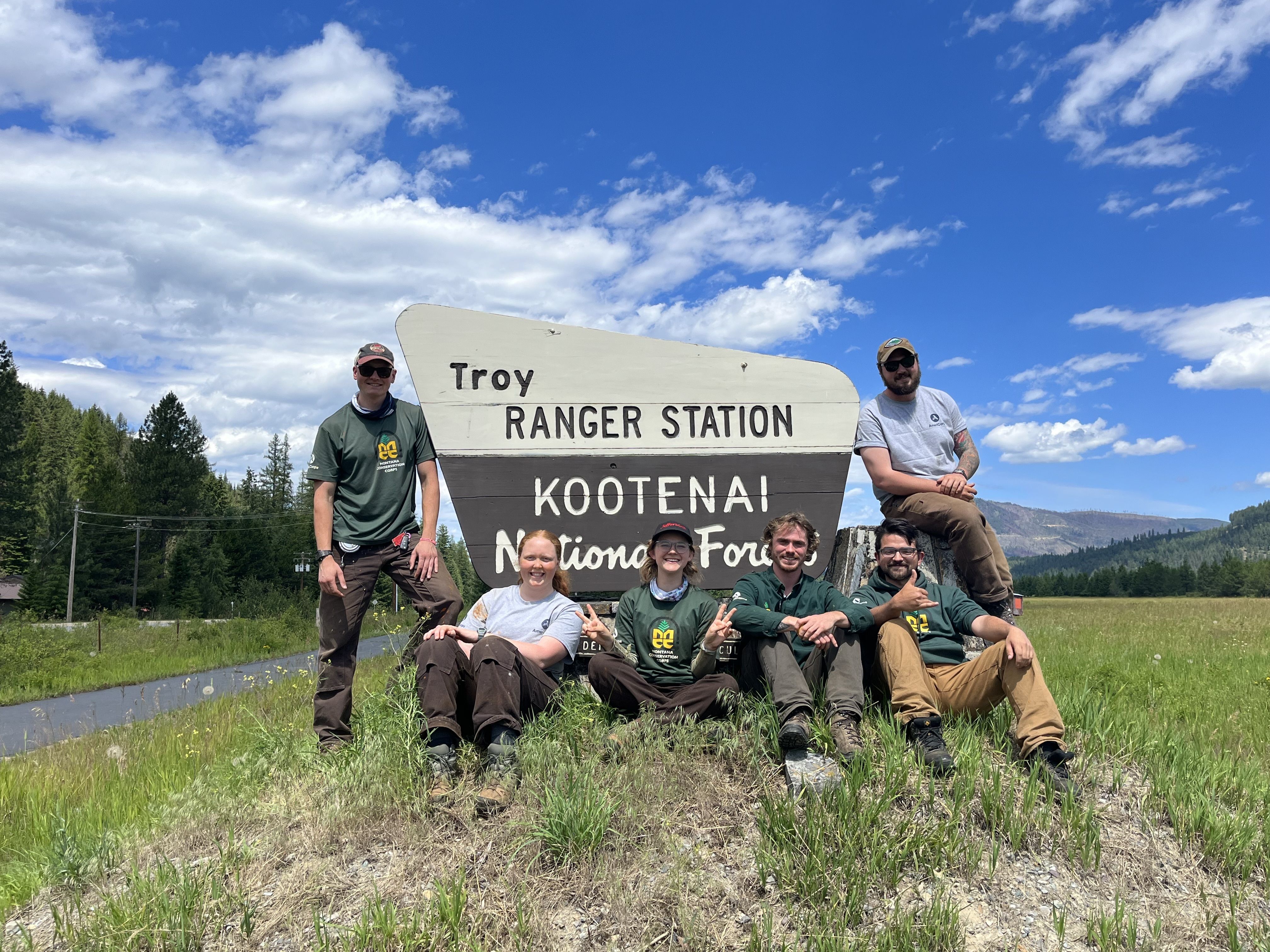 A crew sits and stands around a Forest Service sign that says "Troy Ranger Station, Kootenai National Forest"