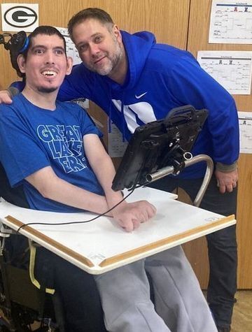 Zach sits in a wheelchair facing the camera while a staff member kneels next to him, also smiling to the camera.