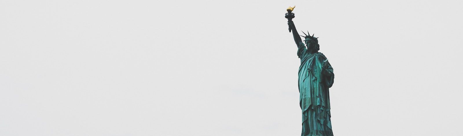Photo of the Statue of Liberty. The background is light grey and the statue is bold teal. The statue stands in the right half of the photo, facing left.