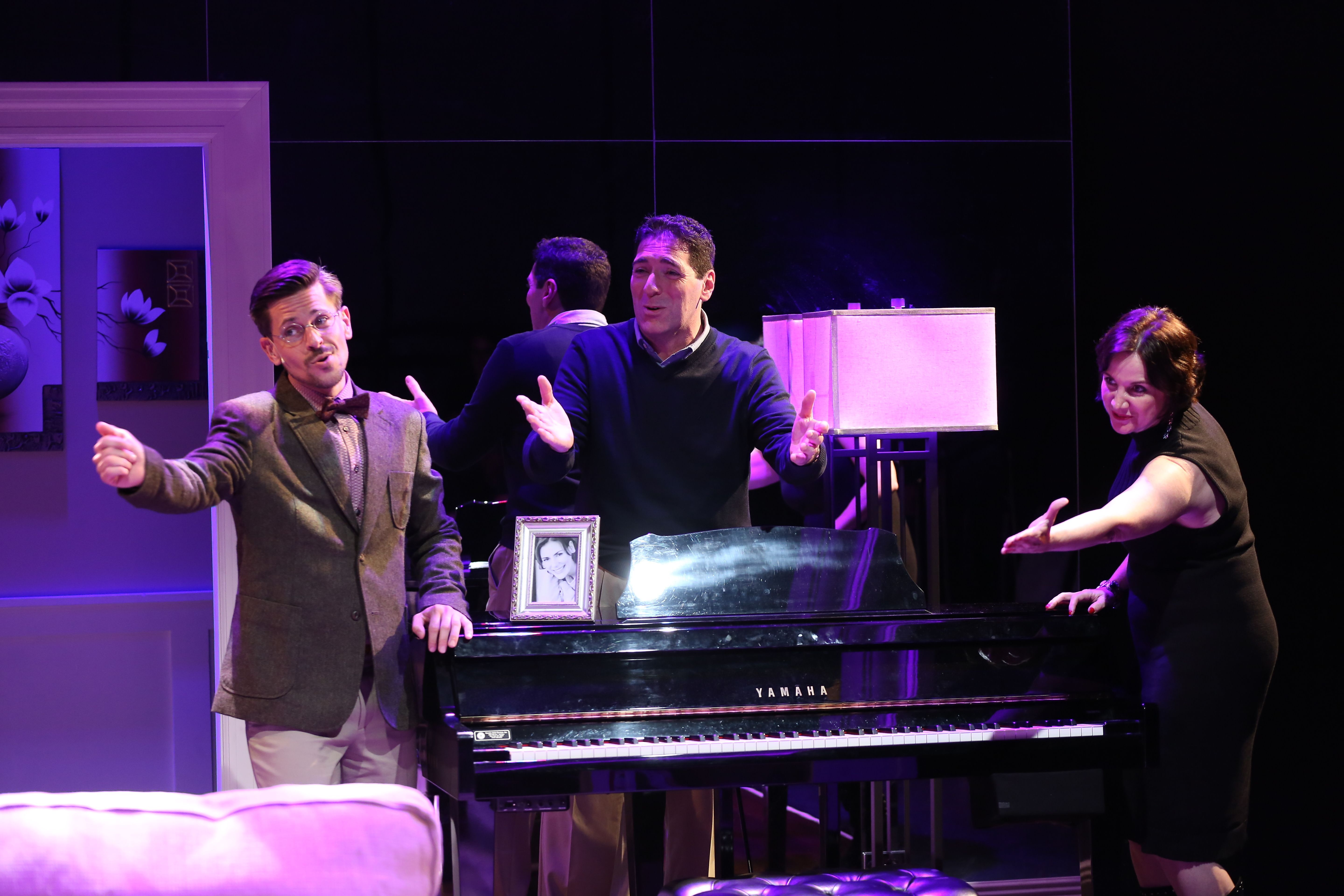 A picture of three actors who’re all standing and performing a scene from the production of The Fourth Wall. They’re all around a piano; two of them are singing, and the third one is gesturing in the same direction.
