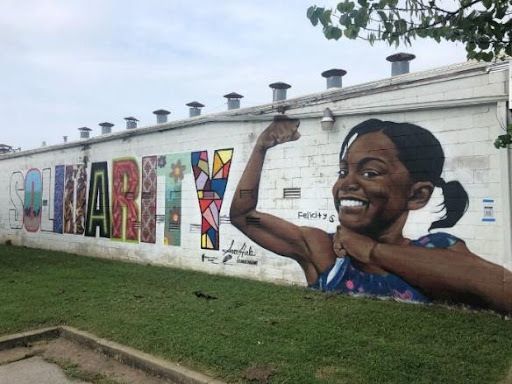 Solidarity: A mural stretches across the side of a white building. Stark block letters spelling out the word Solidarity are filled with different vibrant designs in each letter. A young Black girl in a blue dress is painted at the end of the word, flexing