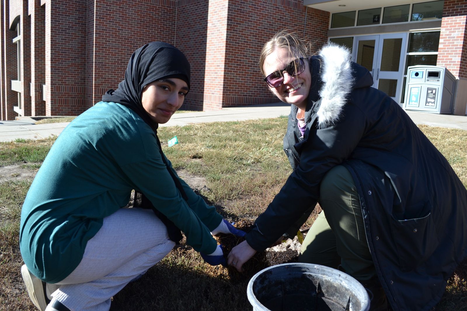 Two women kneel next to a tree they are planting together. 