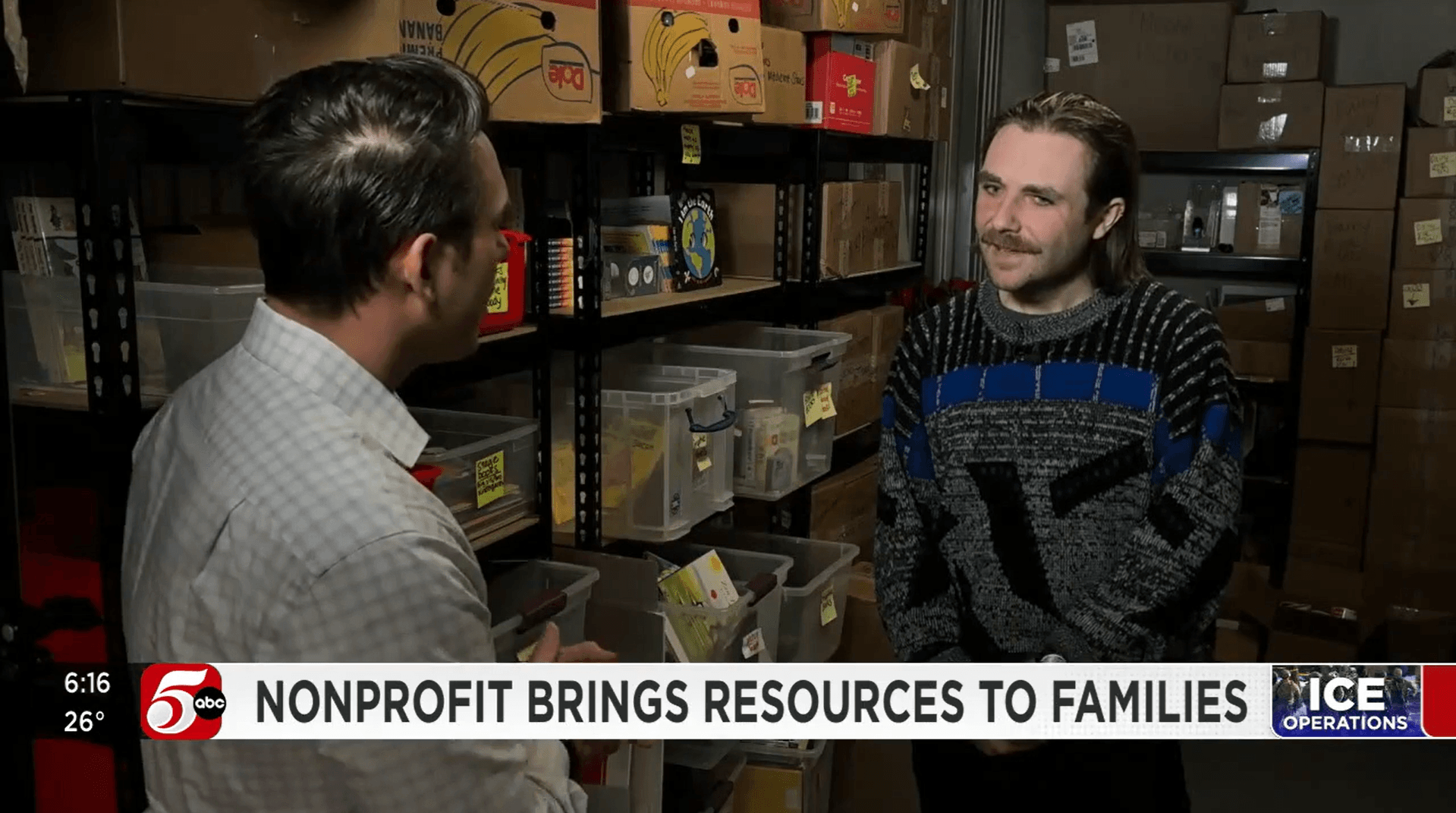 News reporter speaks with man in blue and black sweater in front of shelves of books. 