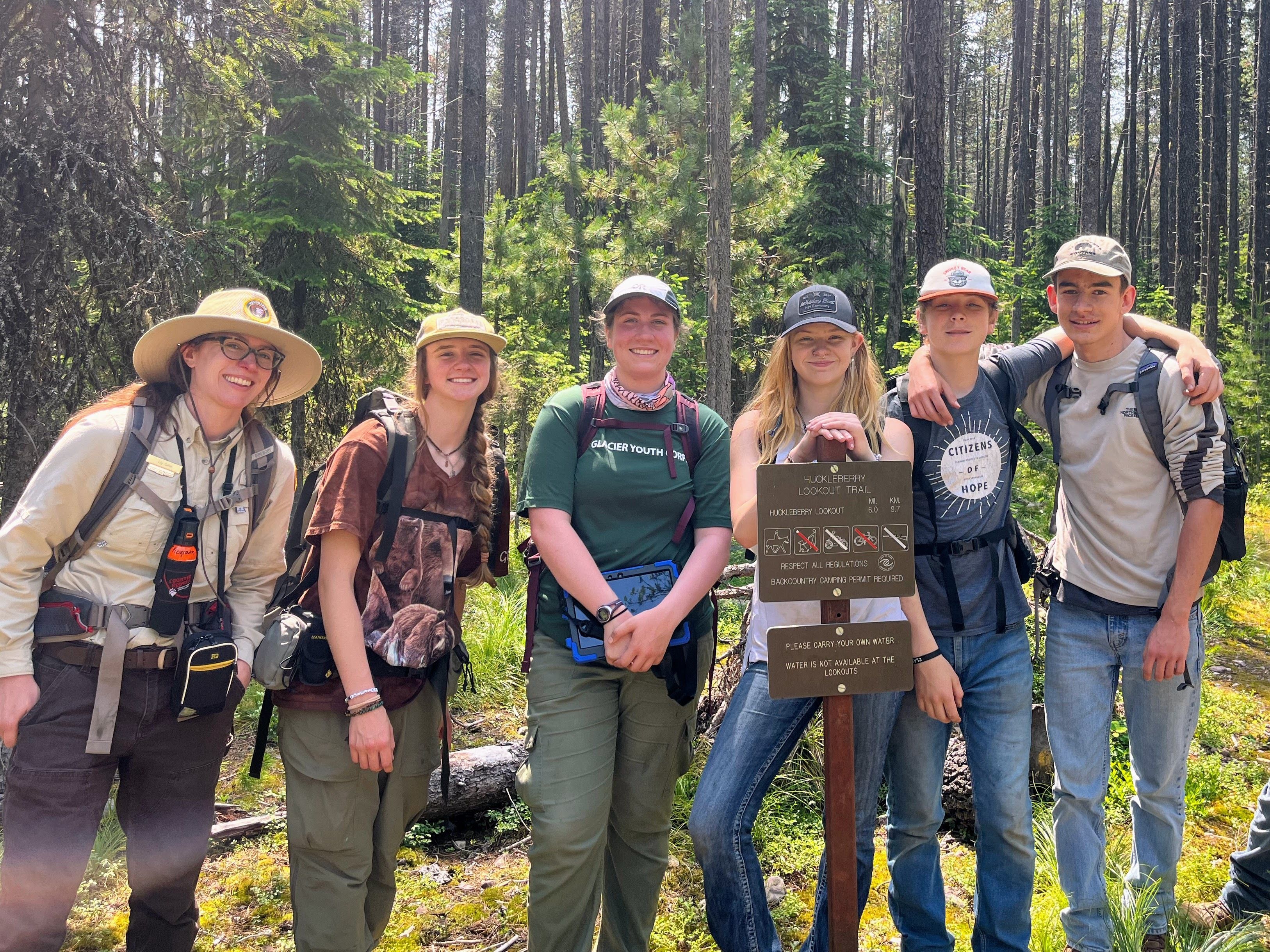 [Image description: Four MCC Members stand with their arms around one another facing the photographer, smiling. Behind them, granulated jagged peaks line the horizon.]