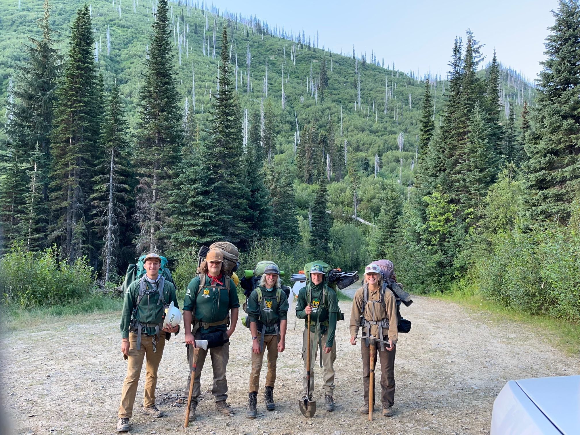 A crew wearing backpacks and gear, stands smiling before a trailhead.