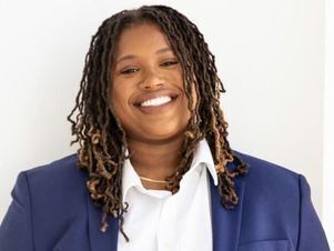 Young Black woman with braided hair parted in the middle and nose ring smiles at camera. She is wearing a blue blazer and white collared shirt.