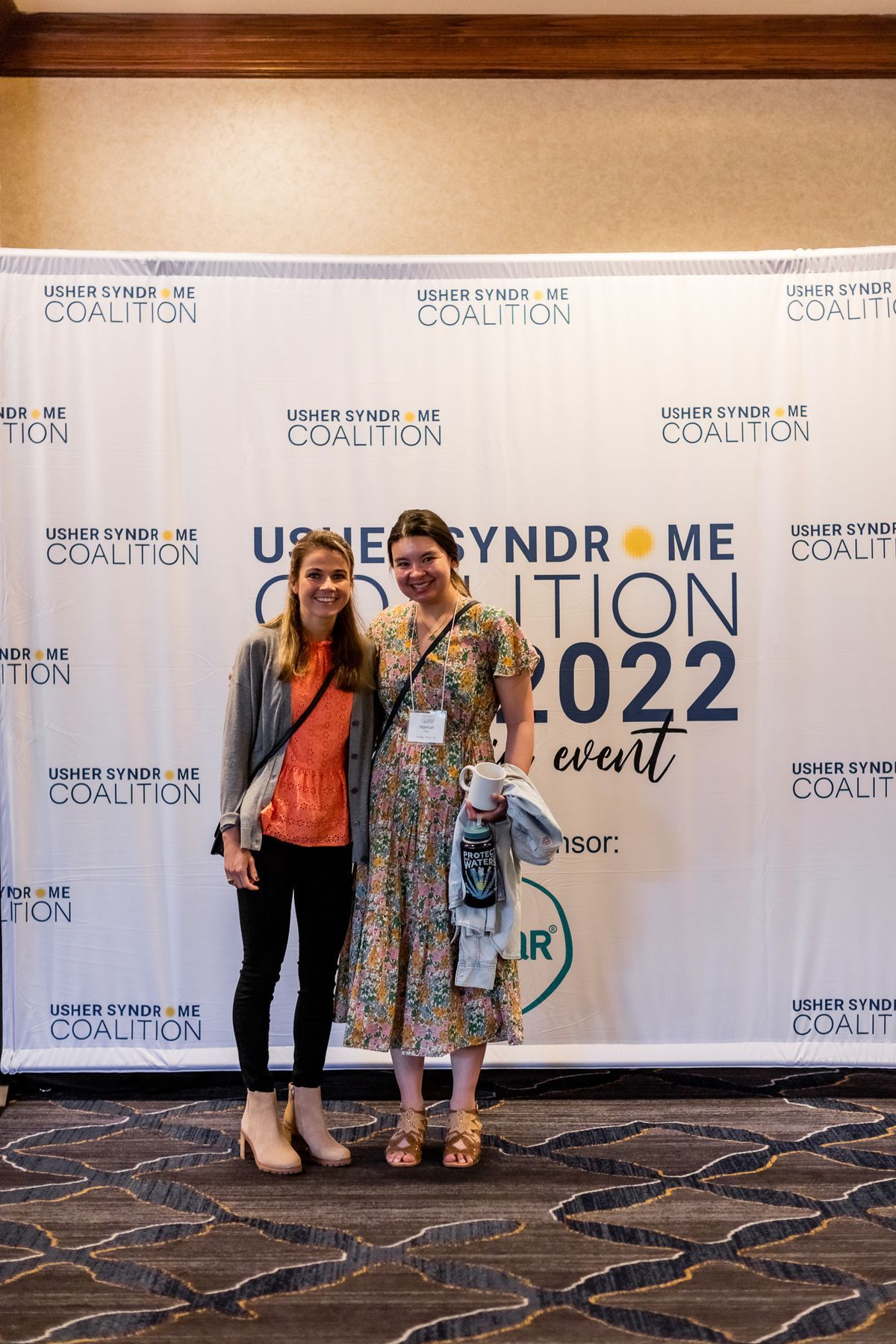 Becca Meyers and Hannah standing together in front of the Usher Syndrome Coalition step and repeat banner.
