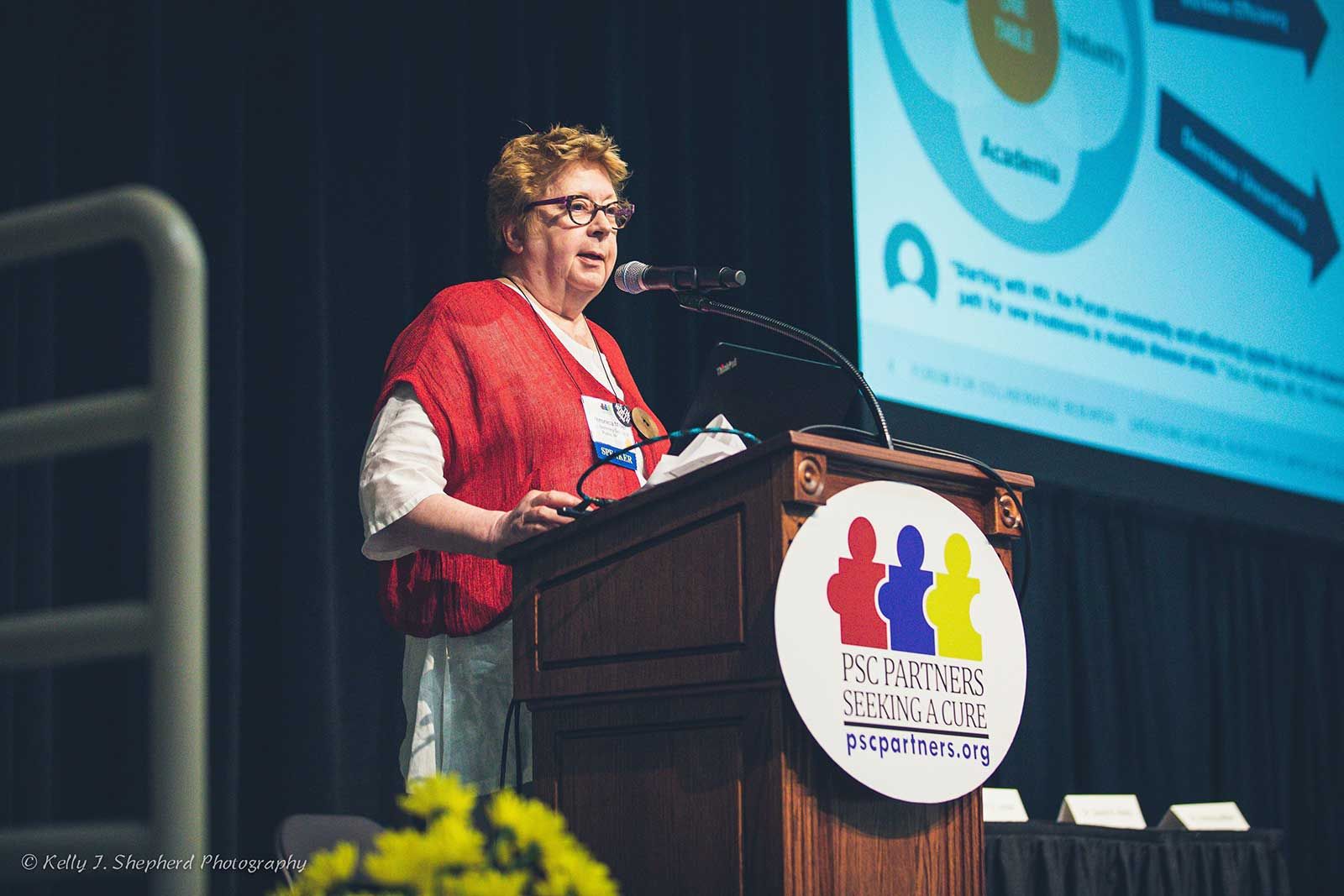 A PSC specialist, Dr. Veronica Miller, stand at a microphone. She is standing a podium with a PSC Partners logo on it. Behind her, you can see a screen showing a presentation.
