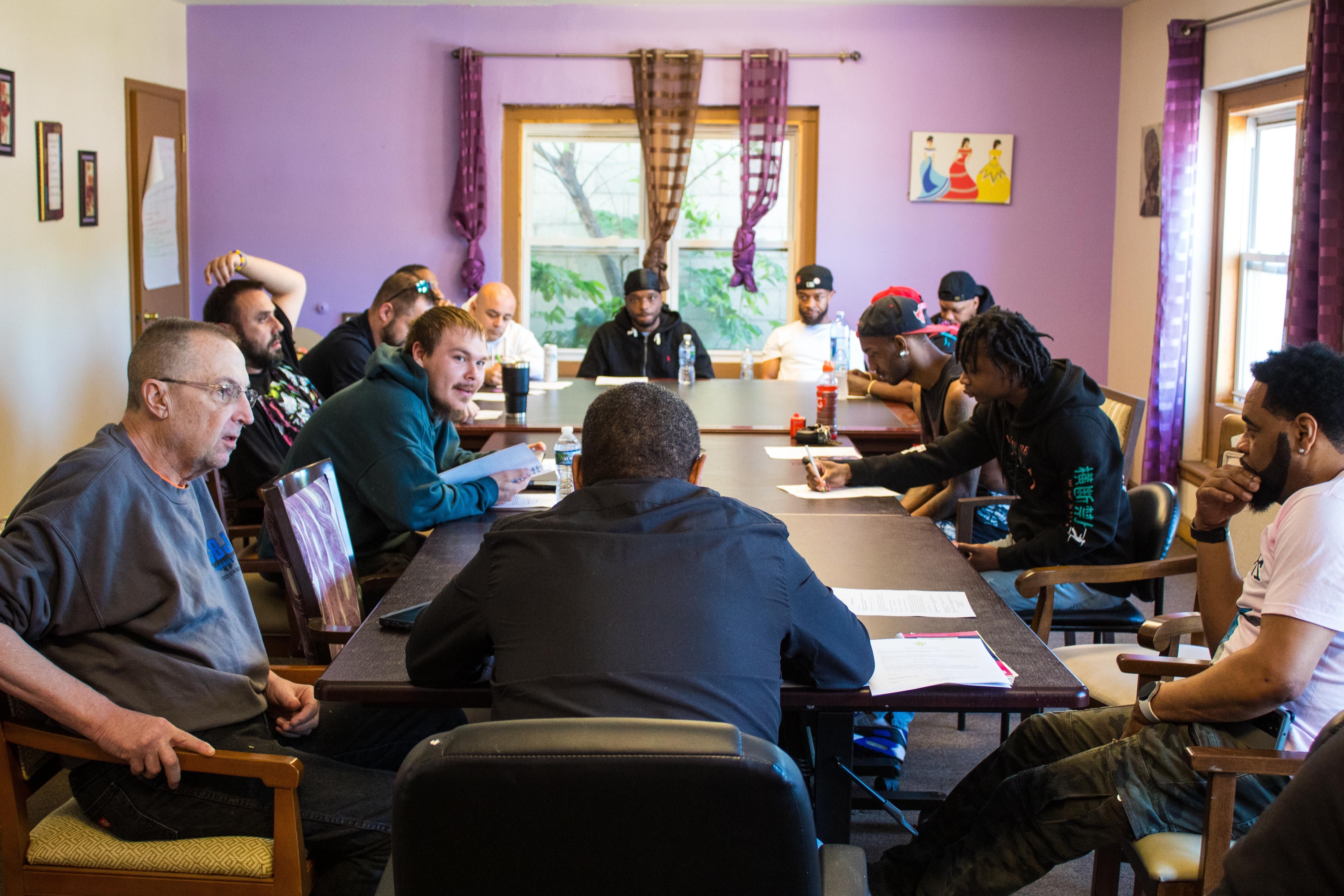 A group of men are sitting around a table in a room with purple walls. They have paper/worksheets in front of them and are engaging in discussion. 