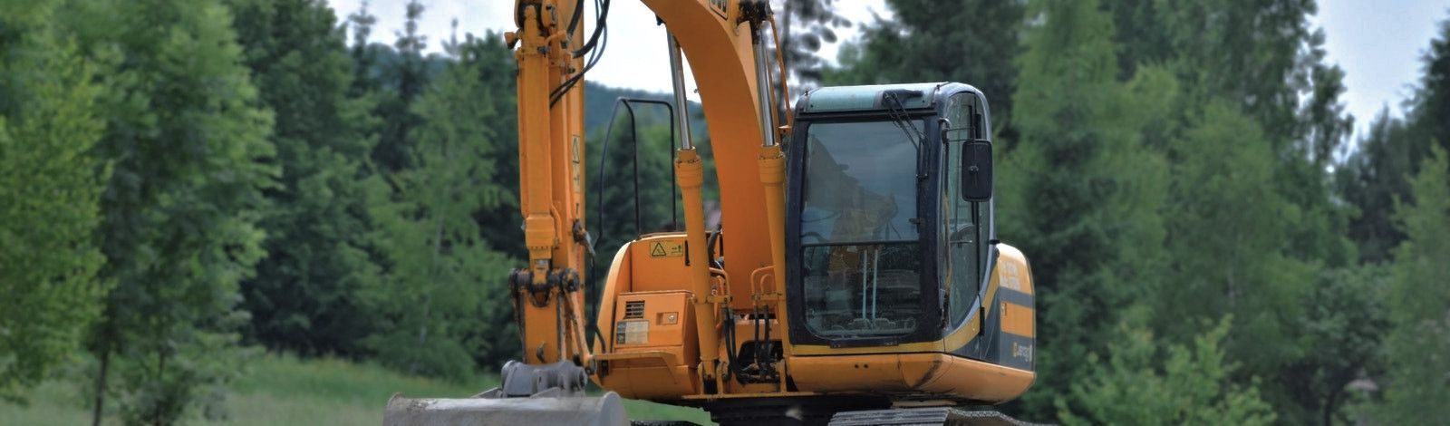 Photo of a large, orange-yellow construction machine with an arm and scoop. The background is a forested area with lots of trees and green grass.