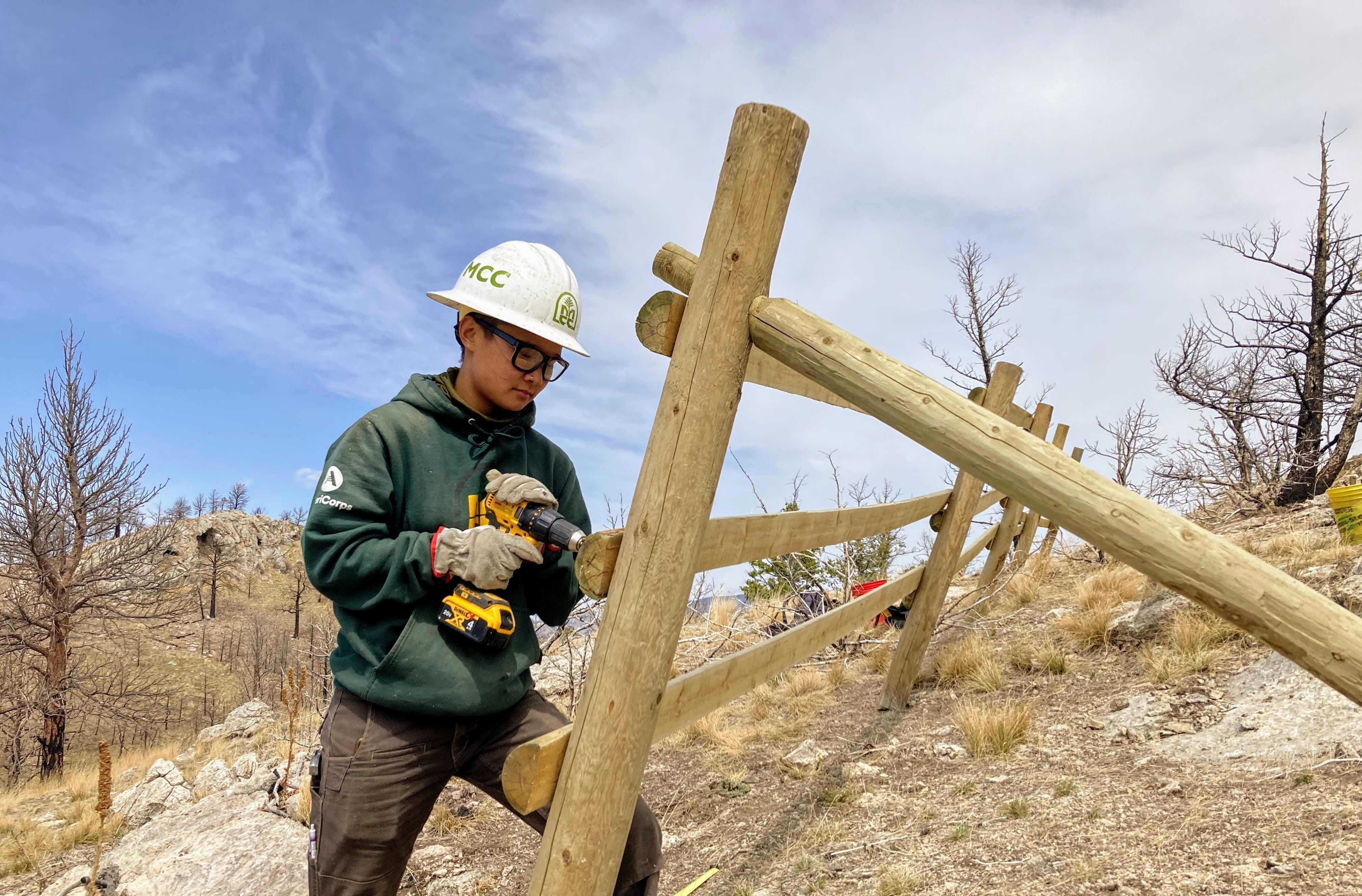 [Image description: A MCC member uses a hand drill to build a fence, wearing their MCC sweatshirt and hard hat.]