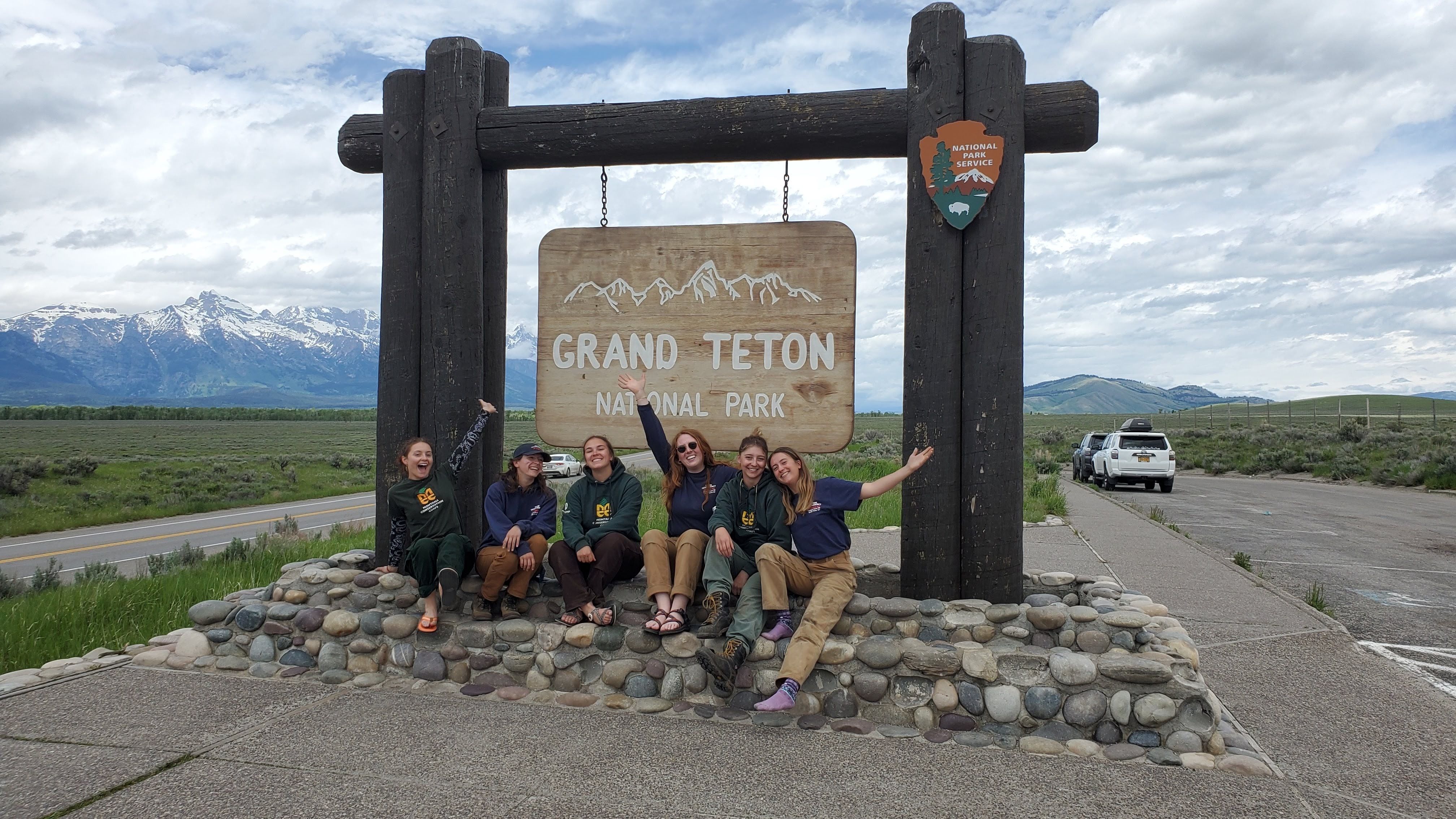 An all women's crew poses in front of the sign for Grand Teton National Park. There are mountains in the background