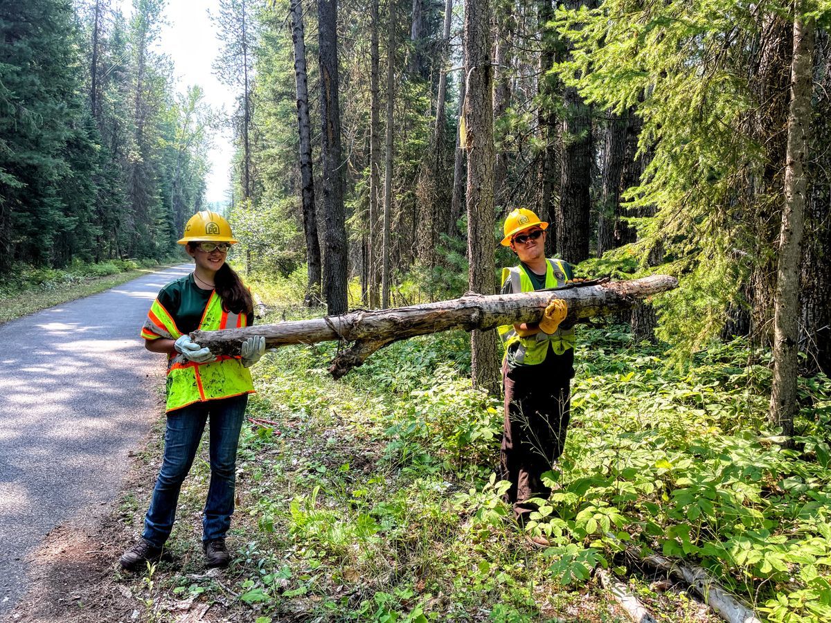 [Image Description: Two MCC Youth members are seen on the side of a small paved trail, hoisting up a fallen log, sporting hard hats, high visibility vests, and glasses.]