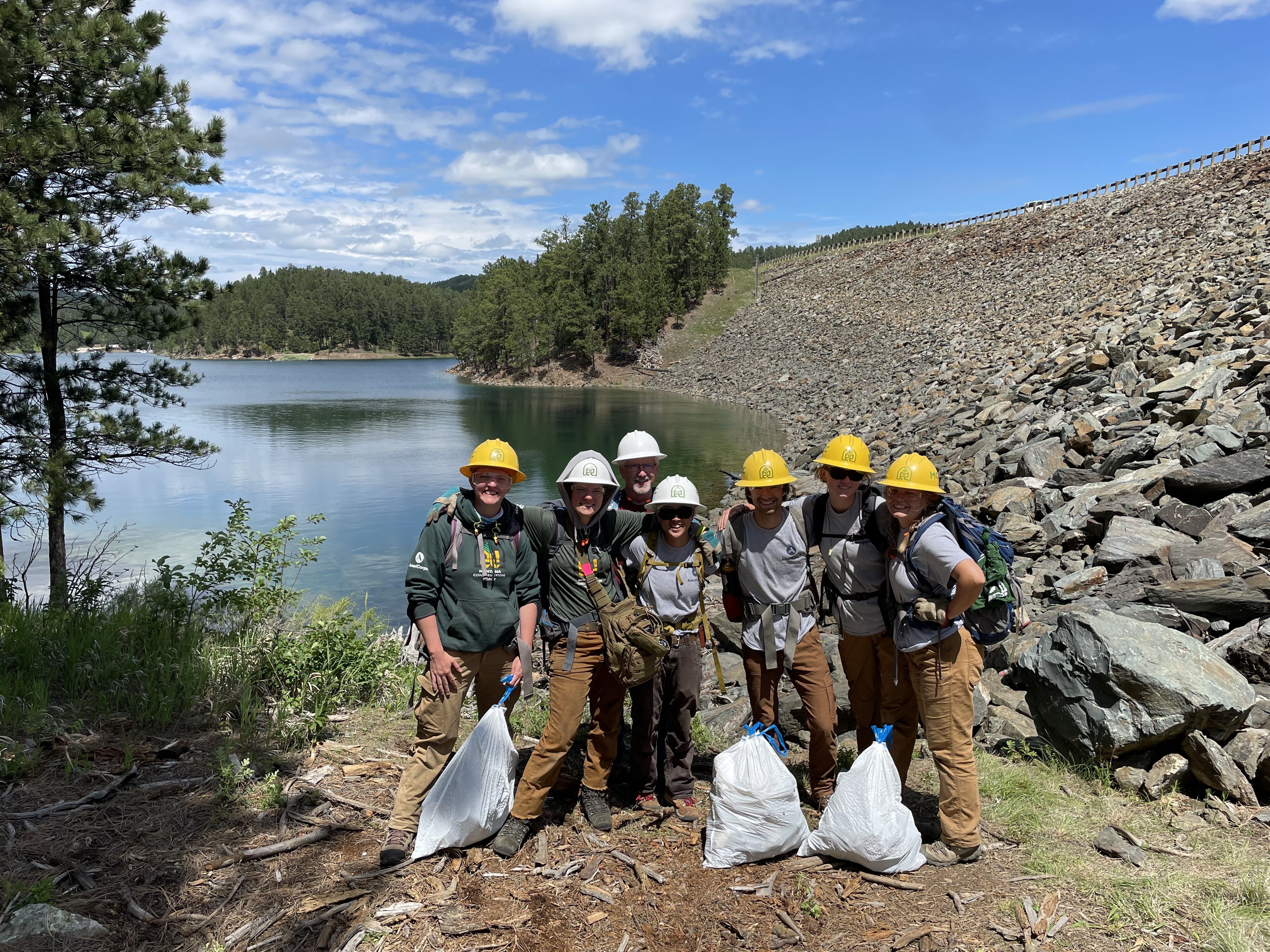 A crew stands on the shoreline of a lake, holding trash bags.