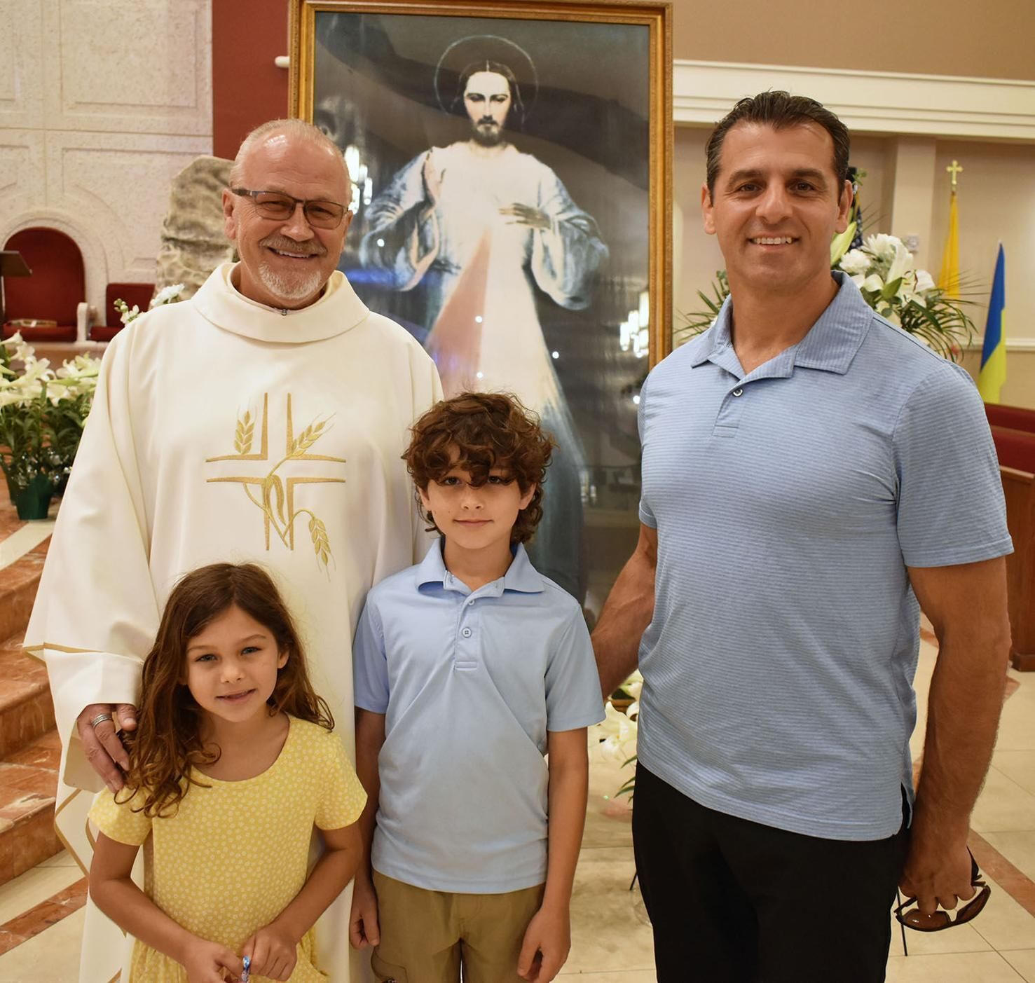 Father Andy Rudnicki, left, pastor of Our Lady Queen of the Apostles Church, stands in front of an image of the Divine Me Otero and his two children April 12, 2026