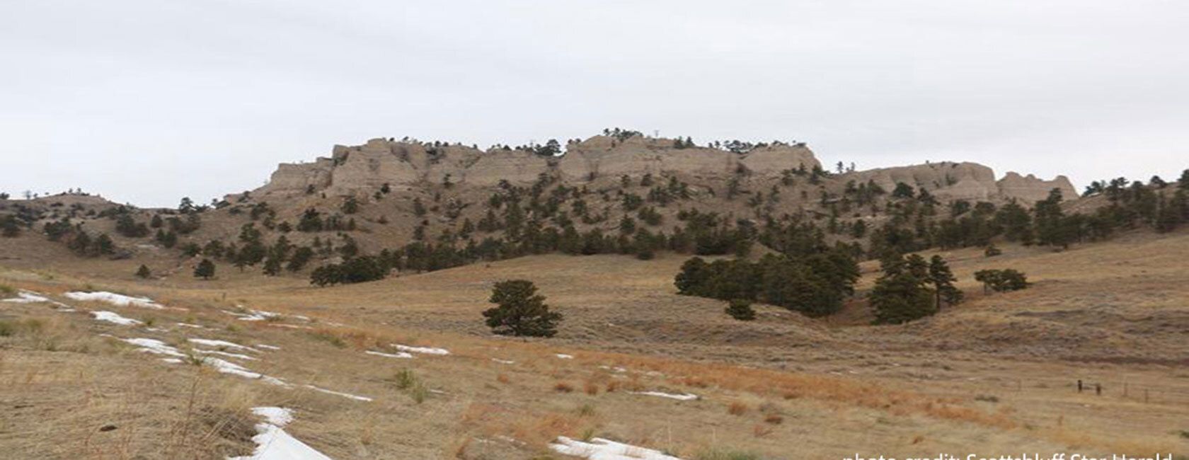Rocky, tree-covered hill under a cloudy sky, with sparse snow patches on a grassy field in the foreground.