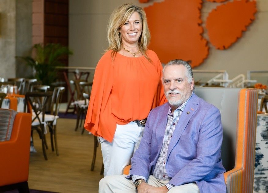 Michael Nieri, owner of Great Southern Homes, is pictured in a blue suit jacket sitting next to his wife Robyn wearing an orange top and white pants. They are posed in front of a tiger paw.