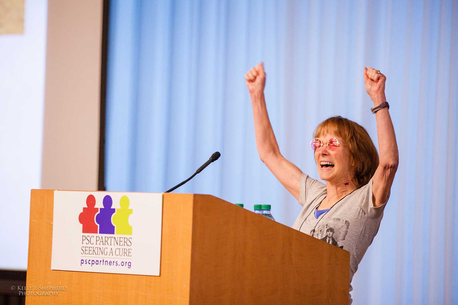 A woman is cheering with her arms in the air. She is standing at a microphone behind a podium that has a PSC Partners logo on the front.