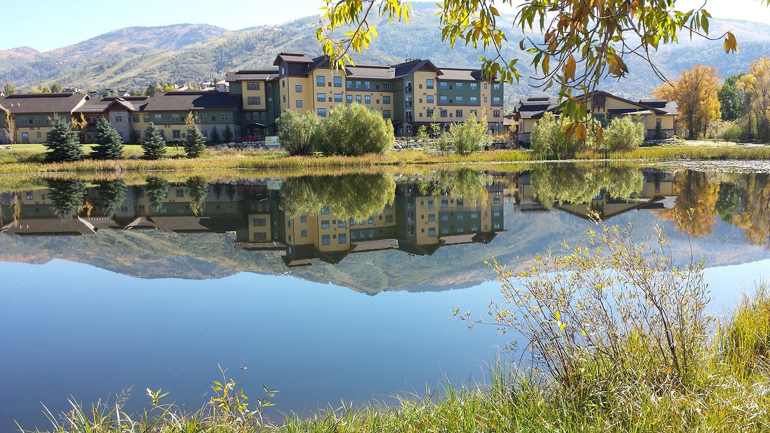 A lakeside building with mountains in the background, reflected in a calm lake under a blue sky.