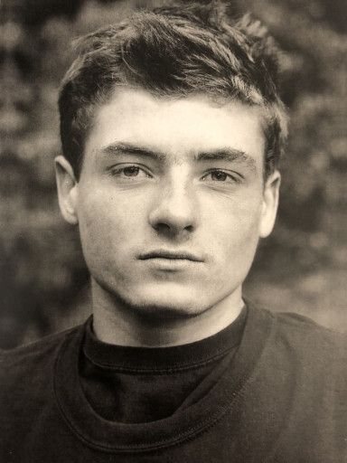 Black and white photo of a young adult with short blonde hair staring at the camera wearing a black t-shirt