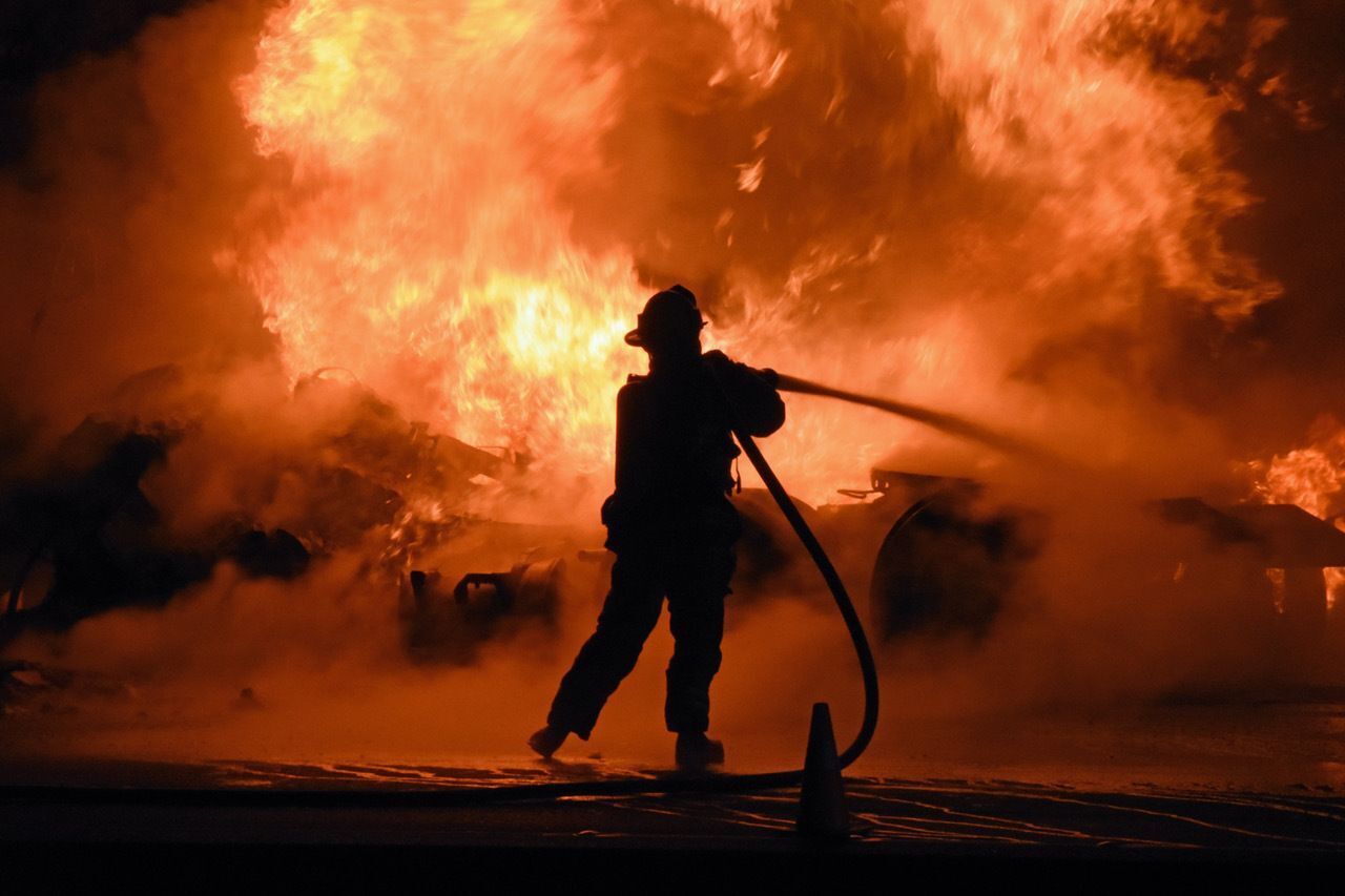 The dark silhouette of a firefighter extinguishing a large vehicle fire using a hose.