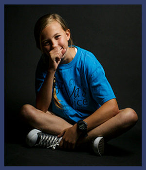 Ava Bullis: portrait of a young girl sitting crosslegged, smiling with her hand covering her mouth.