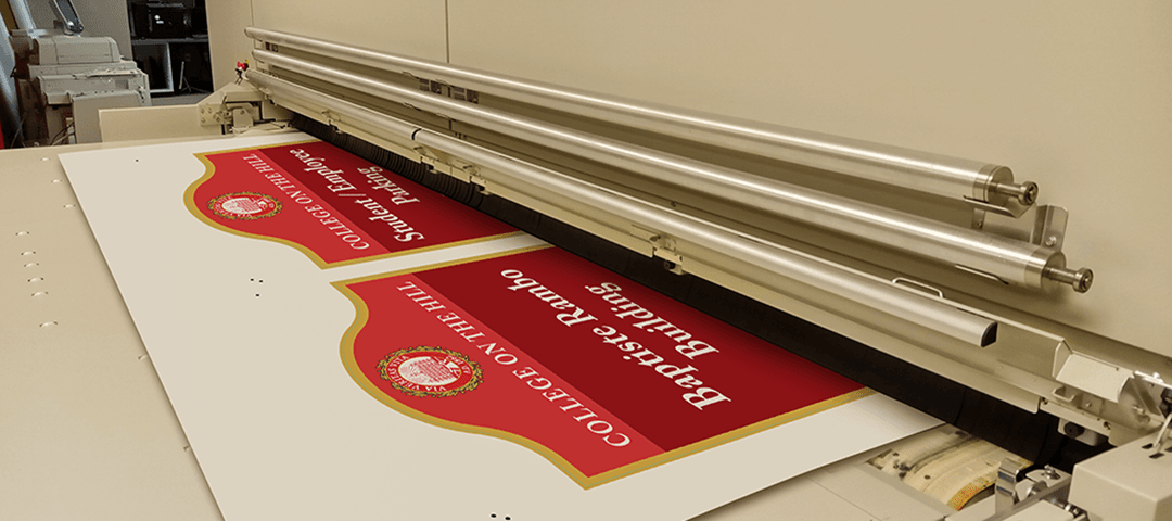 Custom shaped metal signs being printed on large format printer. Signs show College on the Hill seal and text on red background.